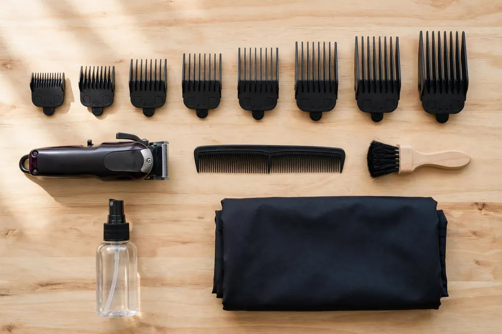 Tools needed to do a fade haircut at home laid out on a table: cordless clipper, full guard set, comb, neck brush, spray bottle and cape